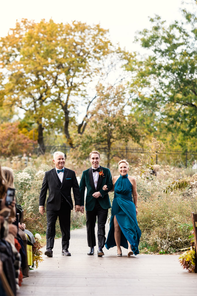 Groom and his parents walk down an outdoor aisle surrounded by greenery and autumn trees during his Lincoln Park Honeycomb wedding ceremony
