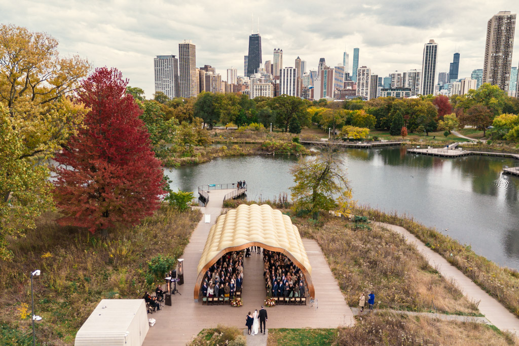 A Cafe Brauer wedding ceremony under an outdoor pavilion with a city skyline and lake in the background.