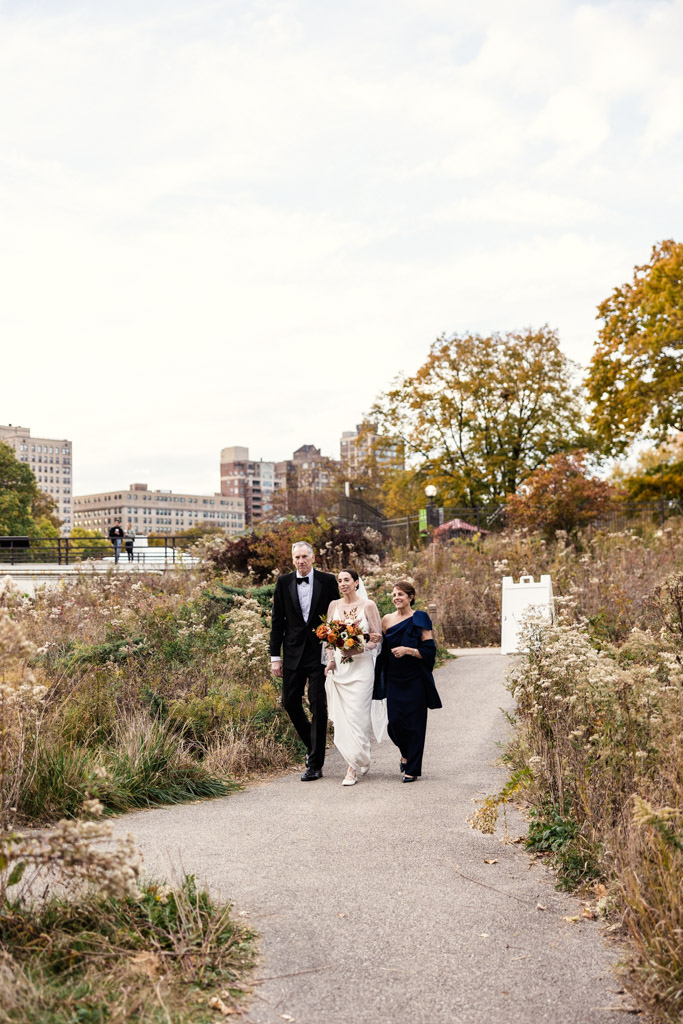 Bride and her parents walks outdoors on a path with autumn trees and city buildings in the background during Lincoln Park Honeycomb wedding ceremony