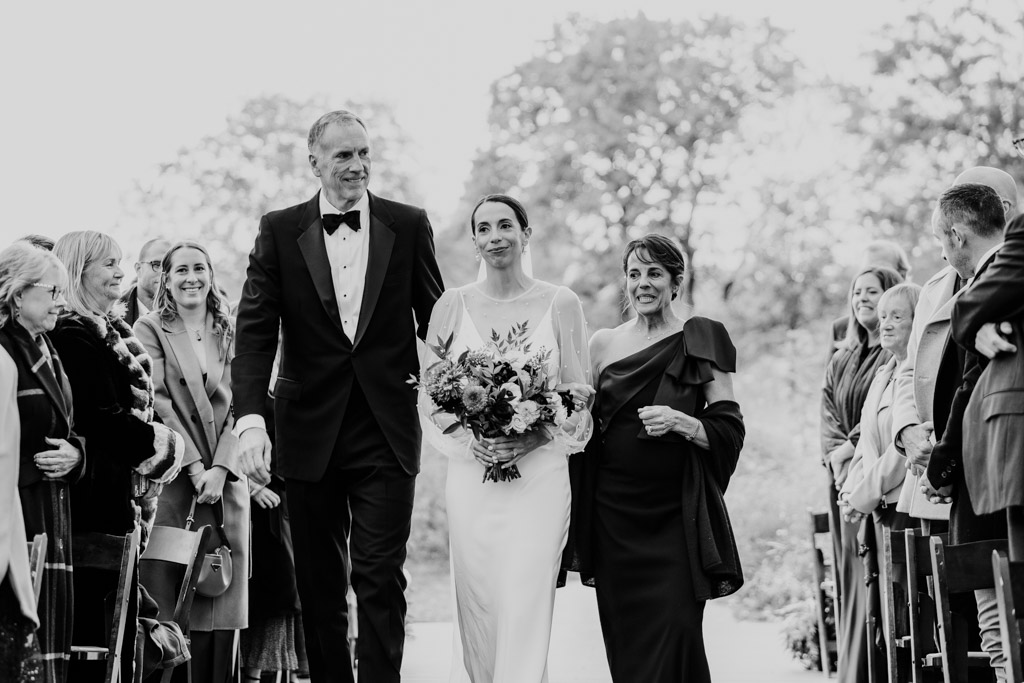 Black and white photo of bride in a white dress walking down the aisle with her parents during her Lincoln Park Honeycomb wedding ceremony, surrounded by smiling guests outdoors