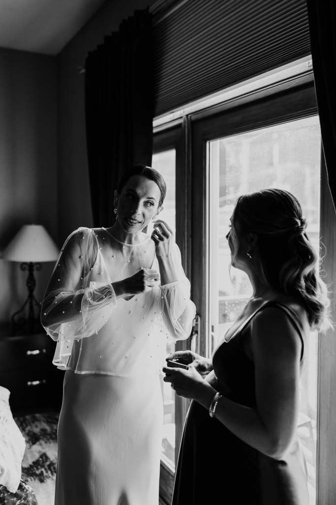 Black and white photo of bride putting on earrings while talking to Maid of Honor by a window in a softly lit room before her wedding at Cafe Brauer