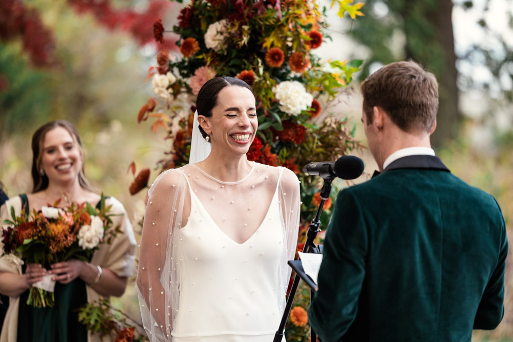 Bride laughs as her groom reads his vows during their Lincoln Park Honeycomb wedding ceremony, surrounded by vibrant autumn flowers