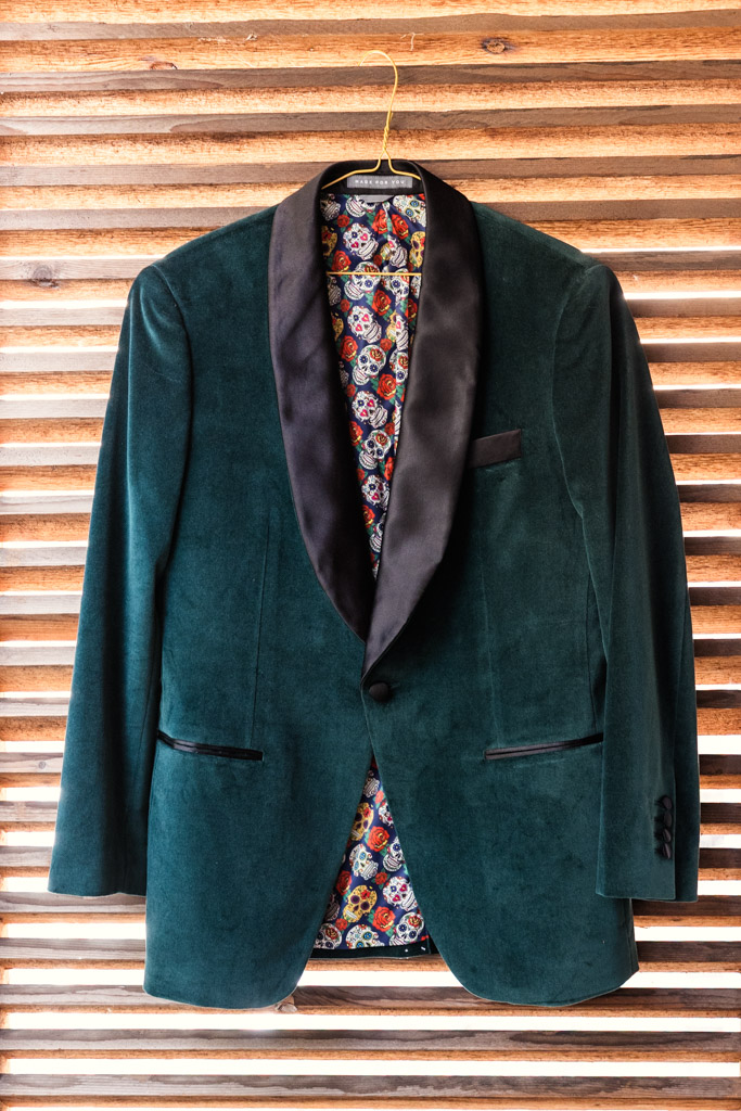 A green velvet tuxedo jacket with black lapels and a colorful skull lining hangs on a wooden slatted wall