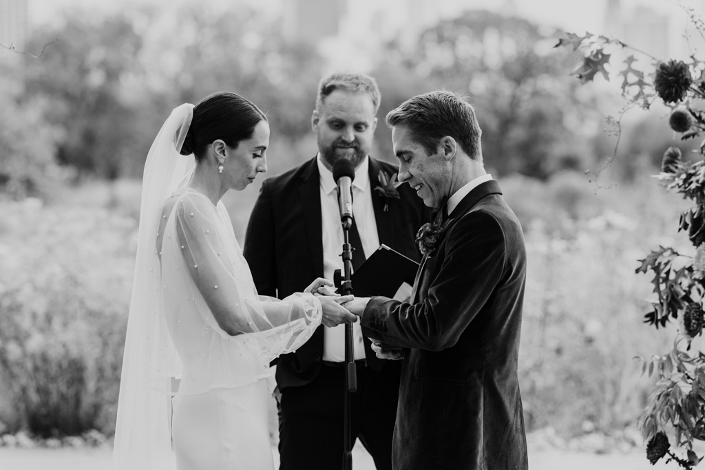 Black and white photo of bride and groom exchanging rings during Lincoln Park Honeycomb wedding ceremony outdoors, with an officiant standing behind them