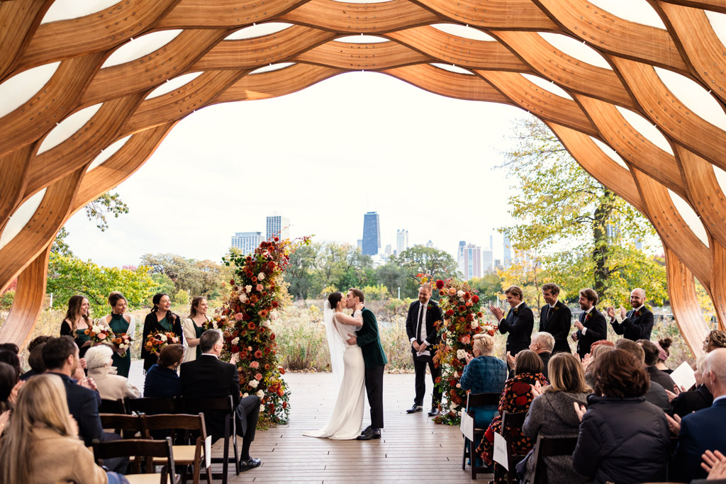 A couple kisses at their outdoor wedding ceremony under the Lincoln Park Honeycomb with guests watching