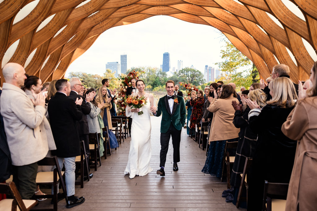 Happy bride and groom walk down the aisle as guests cheer inside the Lincoln Park Honeycomb at their autumn wedding ceremony, with the Chicago skyline behind