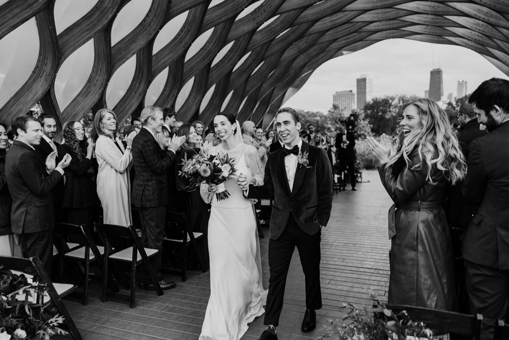 Black and white photo of newlywed couple walking down the aisle after their Lincoln Park Honeycomb wedding ceremony as guests cheer and applaud
