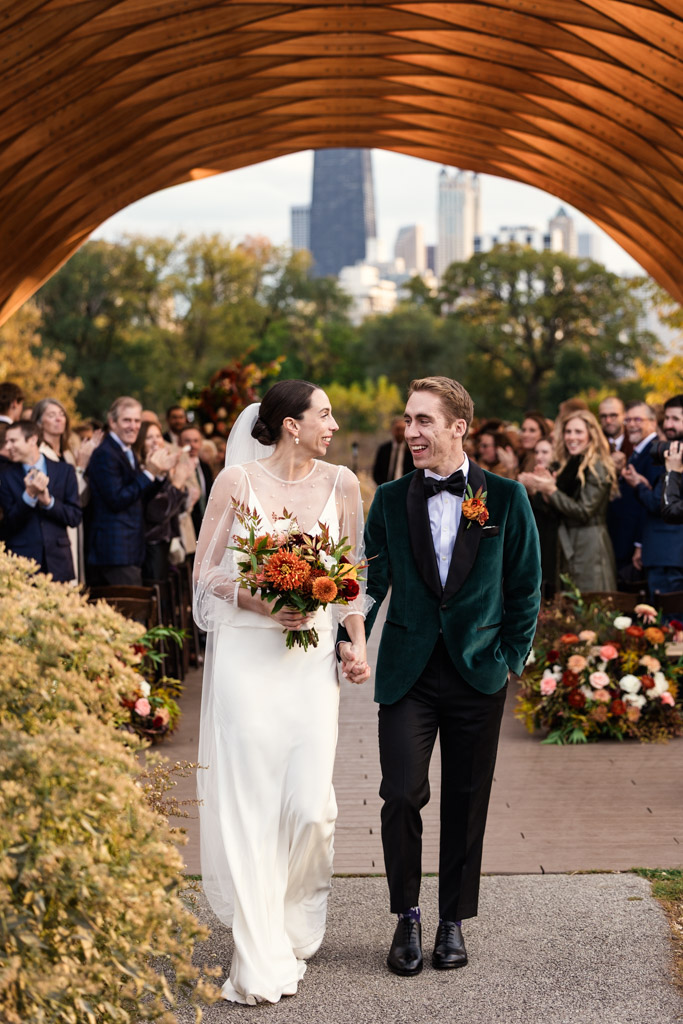 Bride and groom smile while walking down the aisle outdoors at their Lincoln Park Honeycomb wedding ceremony, surrounded by guests and autumn flowers