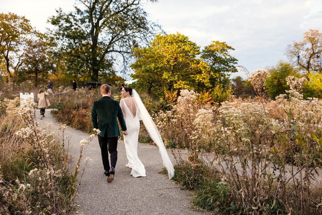 Bride and groom walk hand-in-hand down a path through a field of autumn wildflowers after their Lincoln Park Honeycomb wedding ceremony