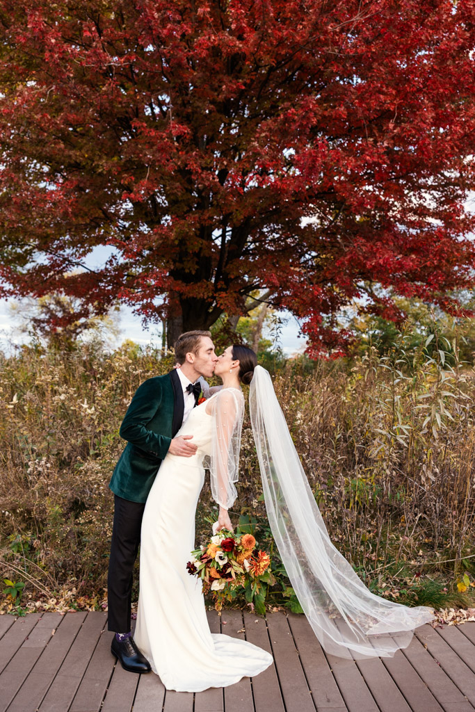 A groom dips and kisses the bride outdoors under a vibrant red tree with autumn foliage before their Cafe Brauer wedding reception