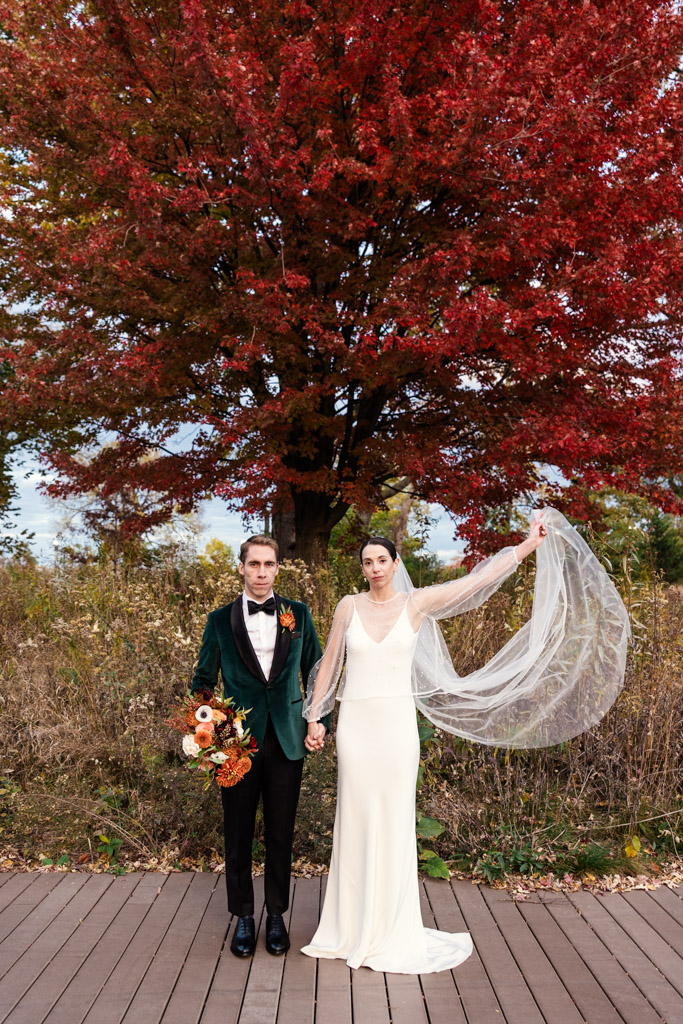 Bride and groom stand under a vibrant red autumn tree at their Cafe Brauer wedding; bride lifts her veil, groom holds bouquet.
