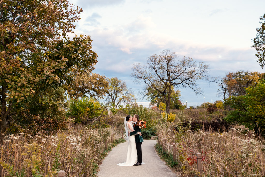 Newlyweds embrace and kiss on a pathway surrounded by autumn trees and tall grasses under a cloudy sky in Lincoln Park