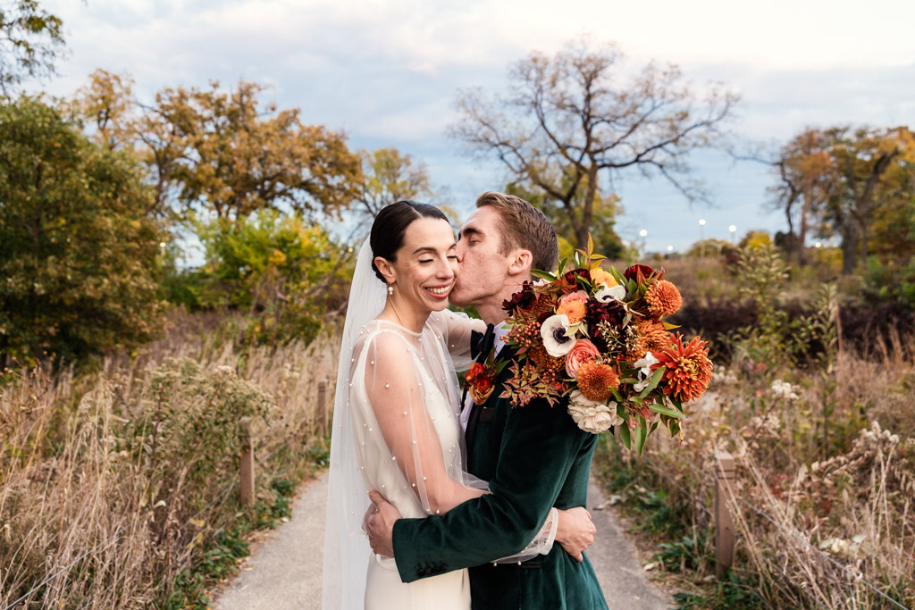 Groom kisses his smiling bride on the cheek as they embrace outdoors in Lincoln Park, the bride holding a colorful bouquet