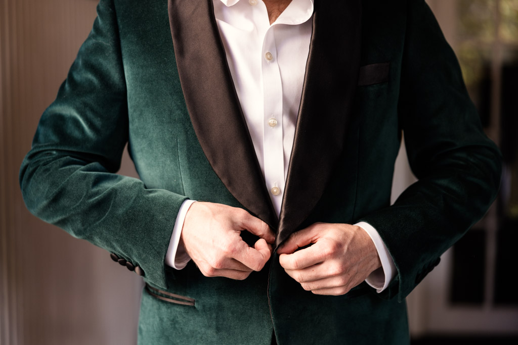 Groom in a green velvet suit jacket buttons it over a white dress shirt, preparing for his Cafe Brauer wedding