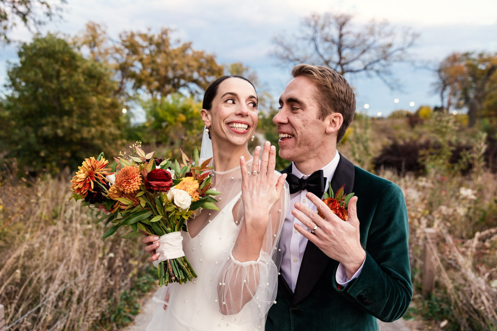 Smiling bride and groom display their wedding rings outdoors at their Cafe Brauer wedding, holding a bouquet with autumn flowers.