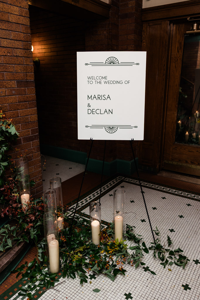 A wedding welcome sign on an easel, surrounded by greenery and candles for Cafe Brauer wedding reception