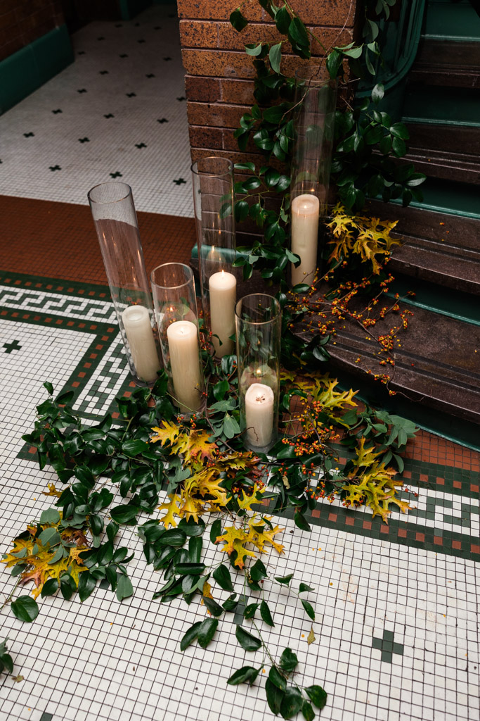 Tall candles in glass vases surrounded by green leaves and yellow flowers on a tiled floor by brick steps for a Cafe Brauer wedding reception