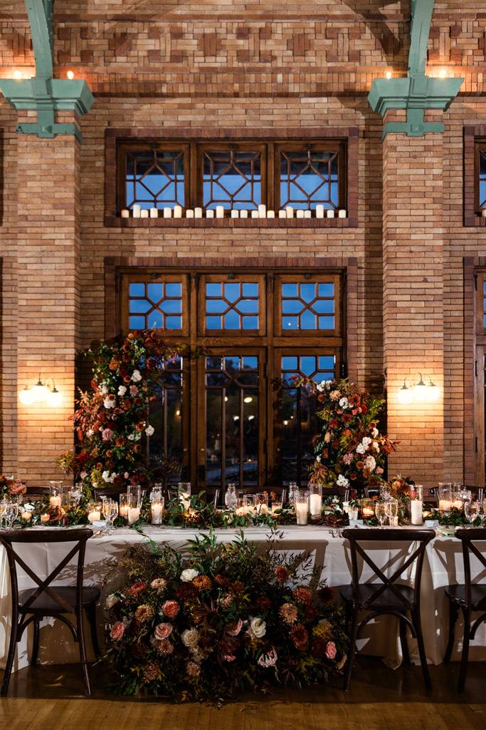 Head table at a Cafe Brauer wedding reception features floral arrangements, candles set against a brick wall