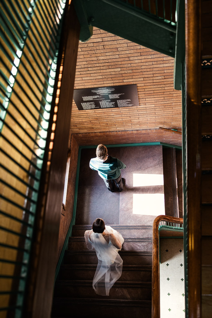 A bride and groom stand on a staircase inside Cafe Brauer during their first look, seen from above, sunlight streaming through a window