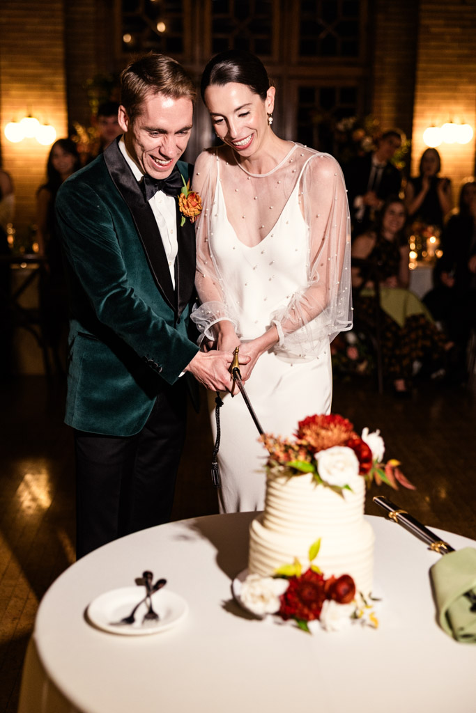 A bride and groom smile while cutting their wedding cake with a sword at their Cafe Brauer wedding reception