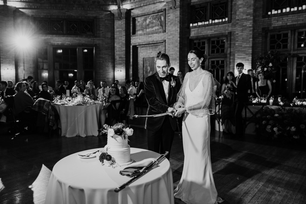 Black and white photo of bride and groom smiling as they cut their wedding cake together with a sword during their Cafe Brauer wedding reception, with guests watching
