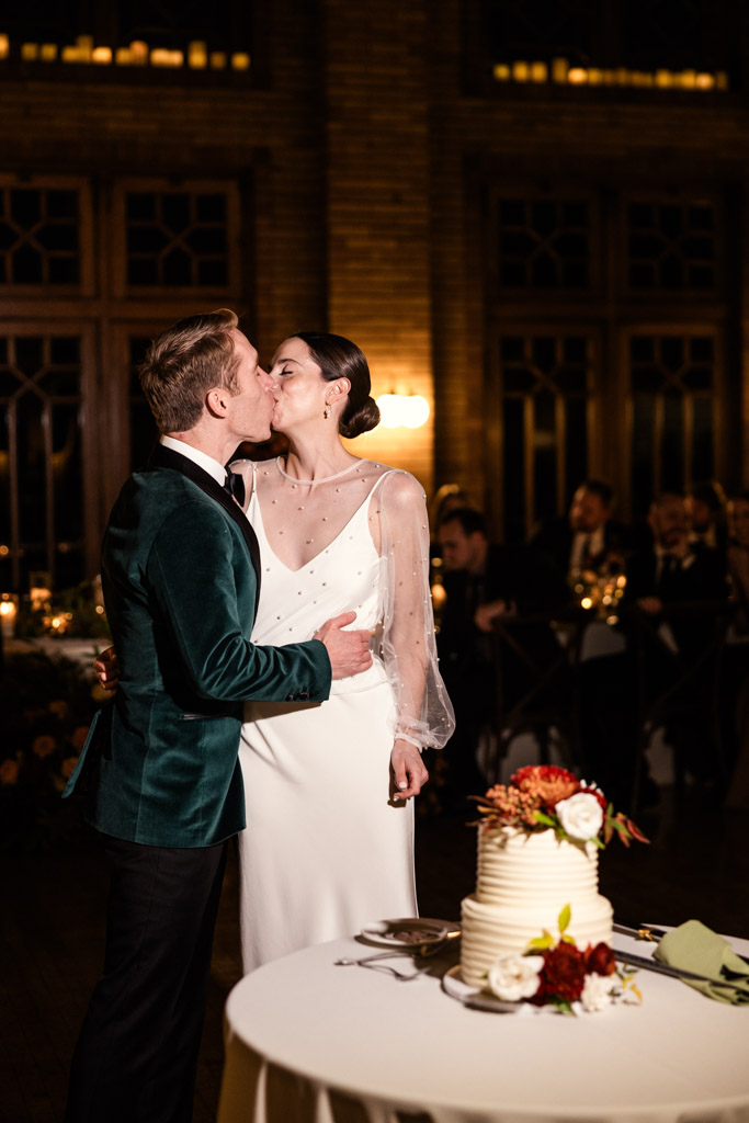 A bride and groom kiss beside their wedding cake at their warmly lit Cafe Brauer wedding reception