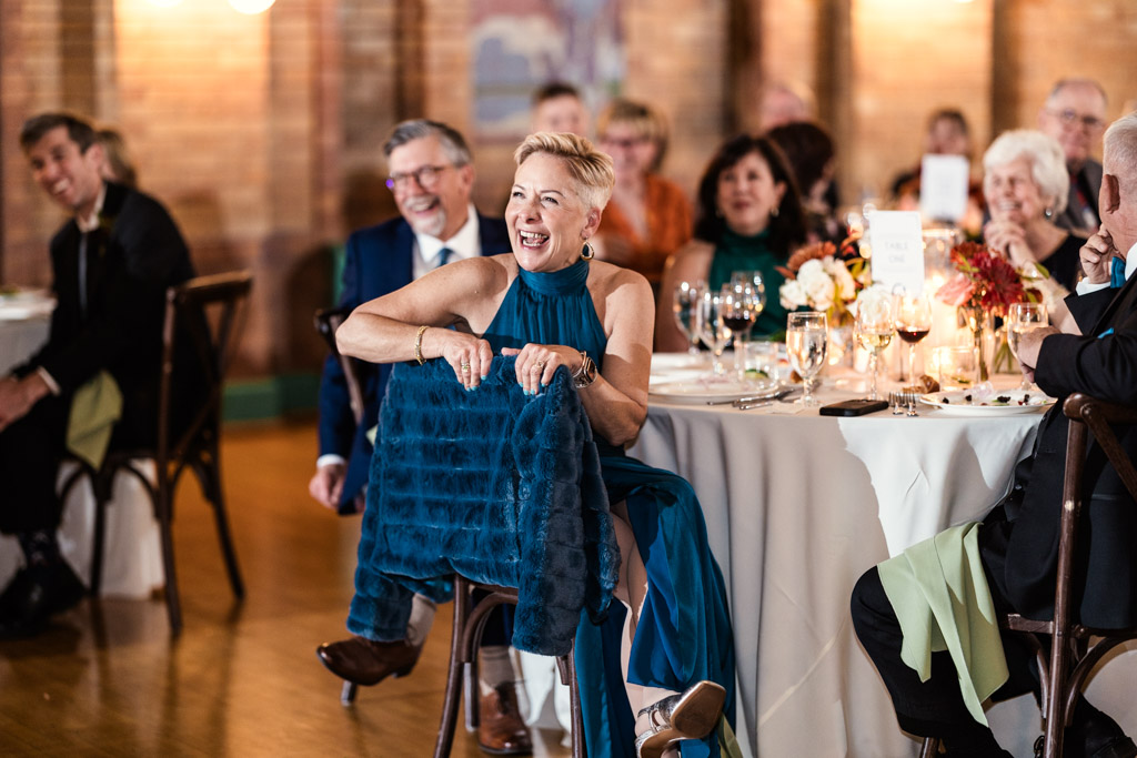 Groom's mother in a blue dress laughs joyfully while seated at a round table during a Cafe Brauer wedding reception