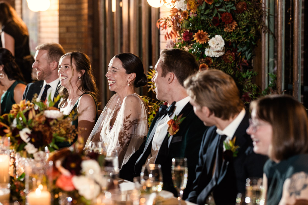 Bride and wedding party members laughing together at the head table adorned with flowers and candles during Cafe Brauer wedding reception