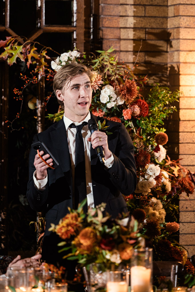 Best Man in a suit gives a speech surrounded by flowers during wedding reception at Cafe Brauer