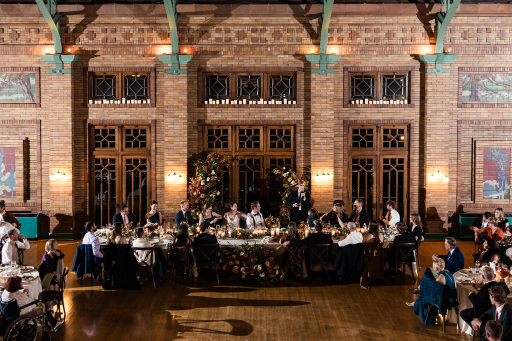Best Man gives speech during Cafe Brauer wedding reception with guests seated at round tables and a flower-decorated head table