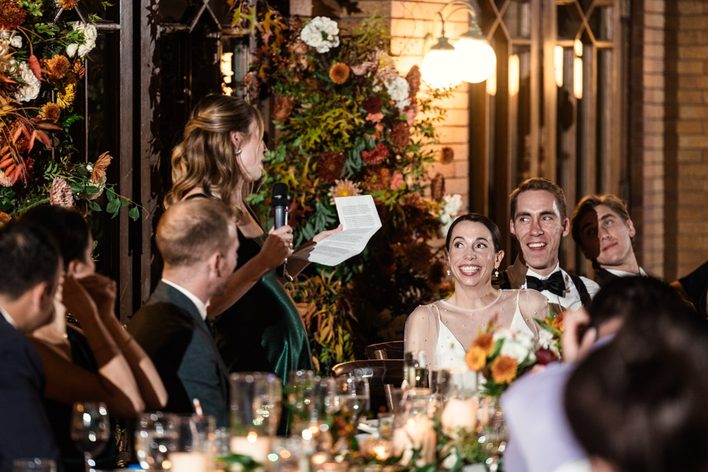 Maid of Honor gives a speech at a Cafe Brauer wedding reception as the bride and groom smile and listen, surrounded by guests and flowers