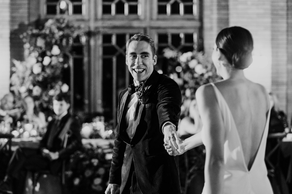 Black and white photo of groom in a tuxedo smiling as he holds hands with his bride in a white dress during their first dance at their Cafe Brauer wedding reception