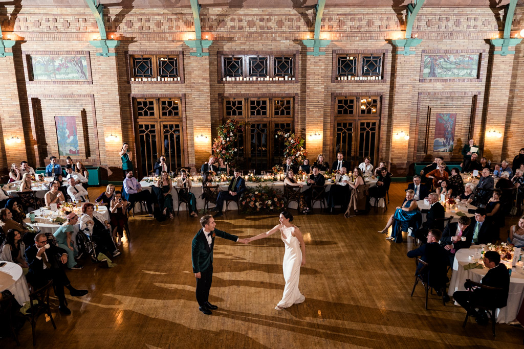 Newlyweds share their first dance in the Cafe Brauer reception space, surrounded by seated wedding guests