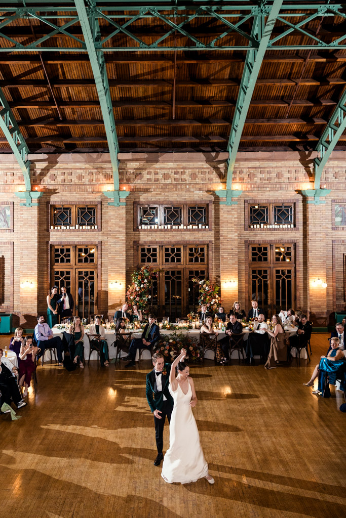 Groom twirls his bride during their first dance inside Cafe Brauer's reception hall, with guests seated around them