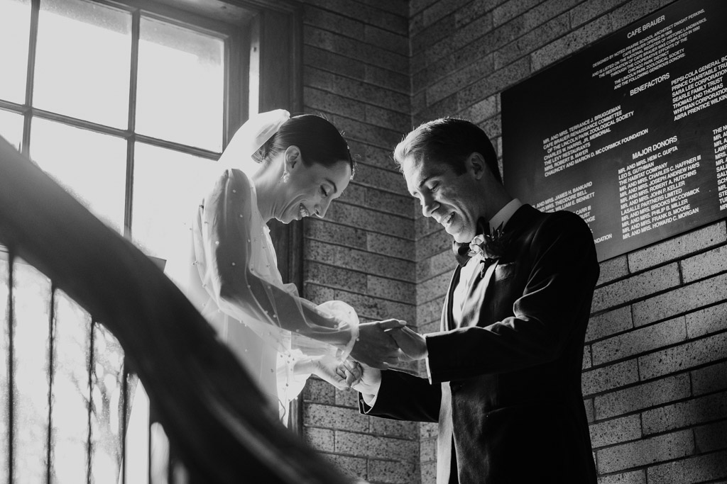Black and white photo of bride and groom holding hands and smiling near a window, bathed in natural light on a staircase at Cafe Brauer