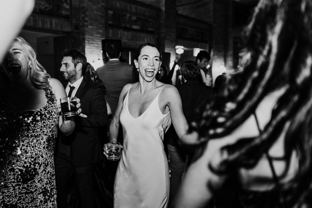 Black and white photo of bride in a white dress smiling and dancng at her Cafe Brauer wedding reception, holding a drink and surrounded by guests