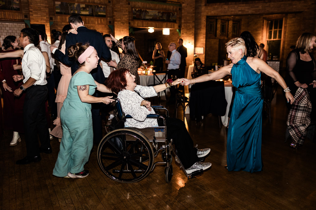 Groom's mother in a teal dress dances with a smiling woman in a wheelchair at Cafe Brauer wedding reception