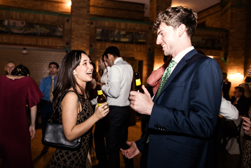 A woman and man smiling and holding beer bottles while talking during an autumn Cafe Brauer wedding.reception