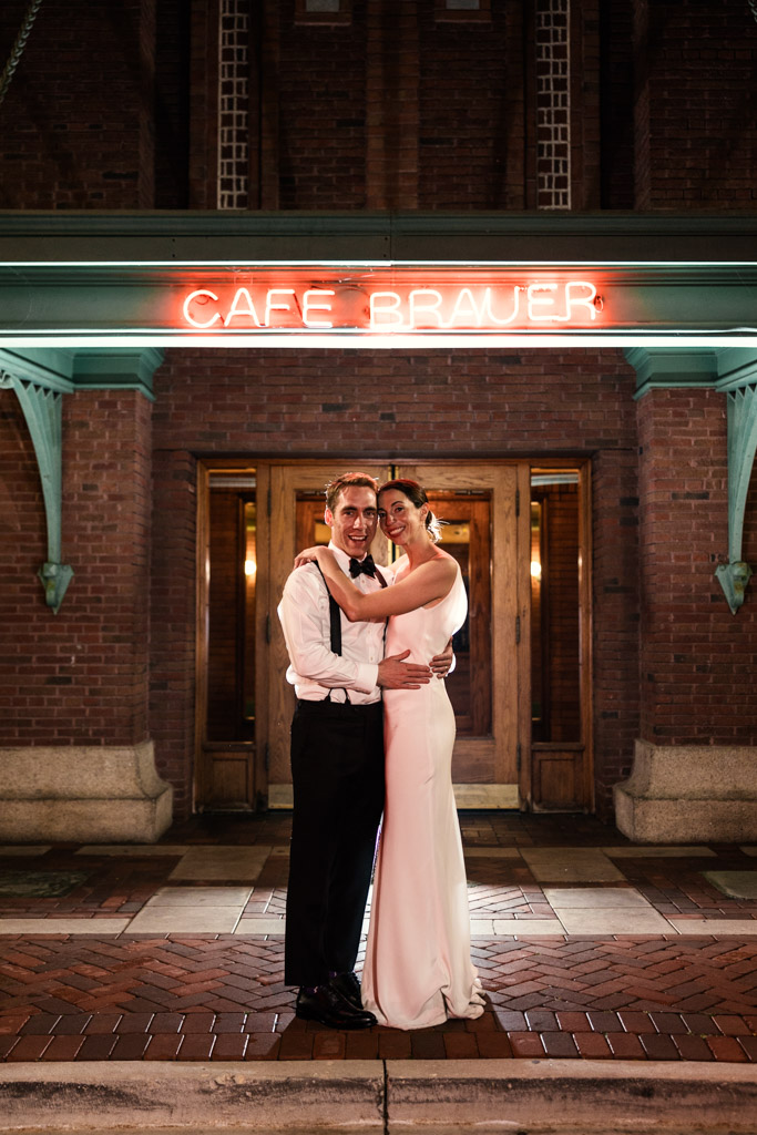 Bride and groom in formal attire embrace at night under a glowing neon 