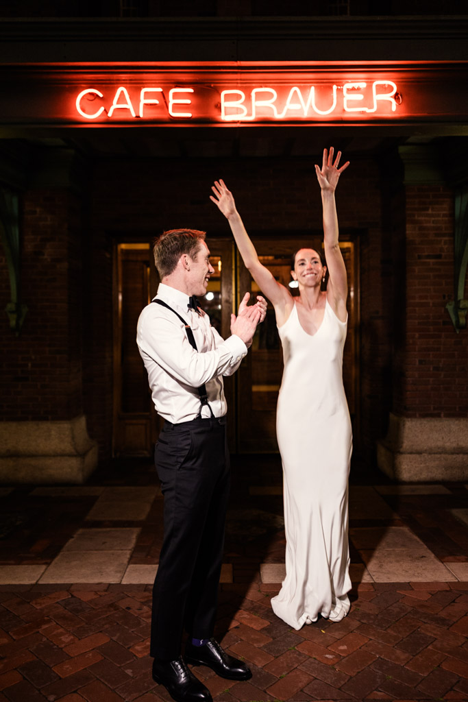 Bride in white dress raises arms joyfully while groom claps outside Cafe Brauer at night under the neon sign