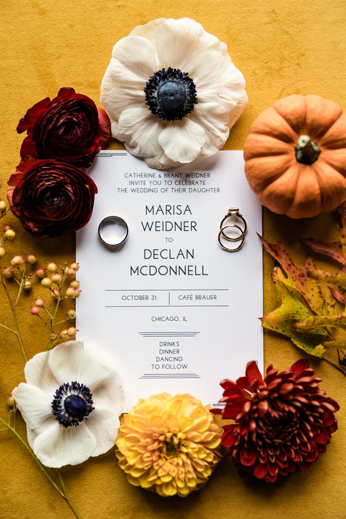 Wedding invitation with rings, flowers, a mini pumpkin, and autumn leaves on a mustard yellow background—perfect inspiration for a romantic Cafe Brauer wedding.