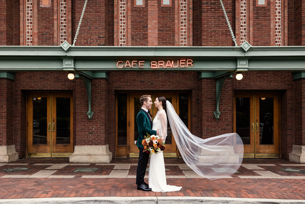 Bride and groom stand facing each other outside Cafe Brauer, her veil flowing in the wind