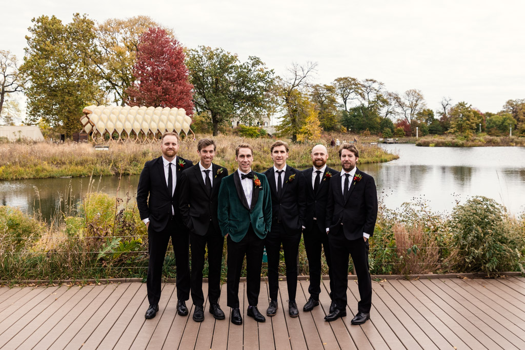 Portrait of groom and groomsmen standing together on a wooden deck by a pond in Lincoln Park, with autumn trees and foliage in the background