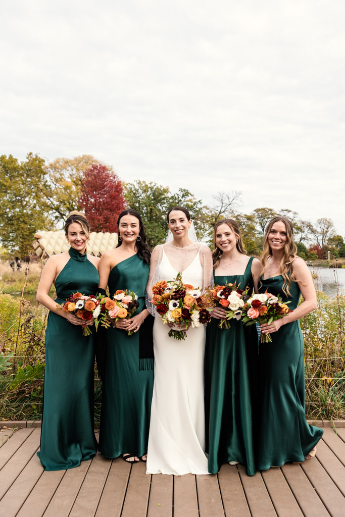 A bride in white stands outdoors in Lincoln Park with four bridesmaids in green dresses, all holding flower bouquets