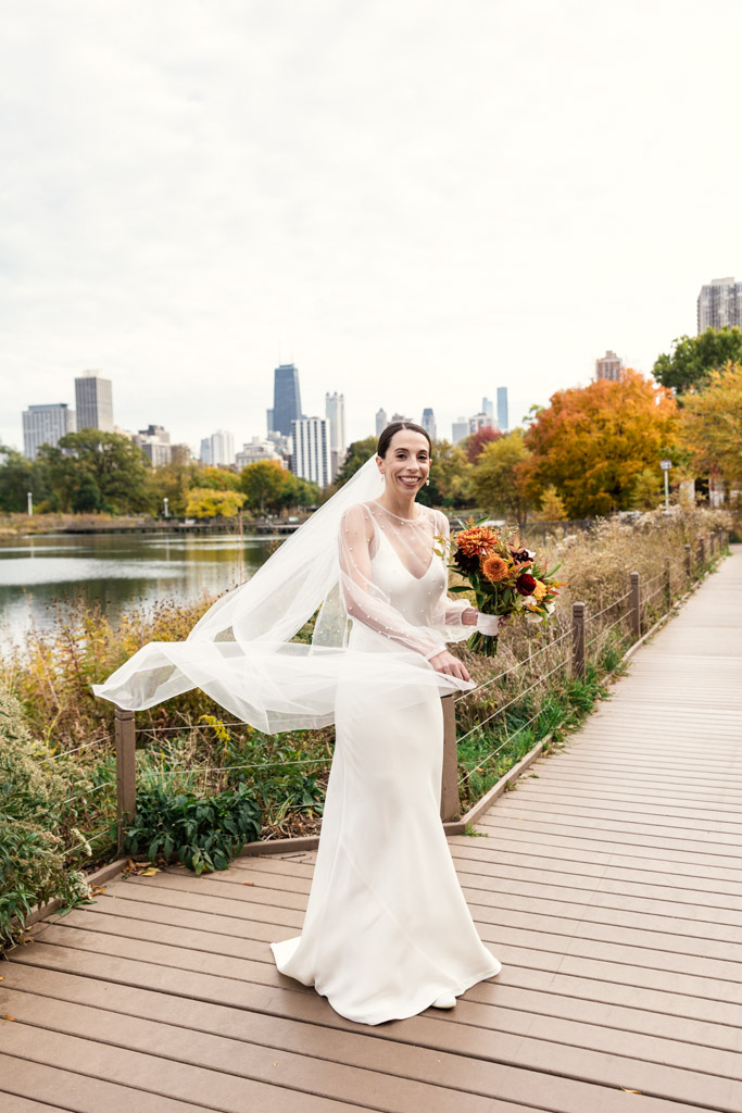 A bride in a white dress holds a bouquet on a Lincoln Park boardwalk with downtown Chicago buildings and autumn trees behind her before her Cafe Brauer wedding