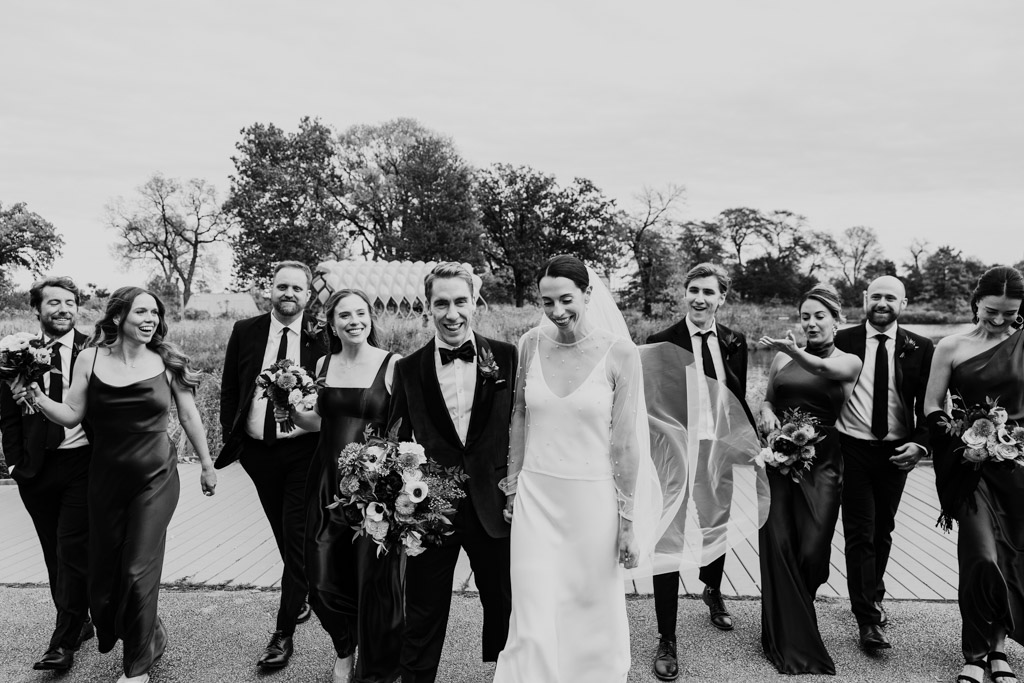 Black and white photo of happy bride and groom walking outside Cafe Brauer surrounded by their wedding party in formal attire