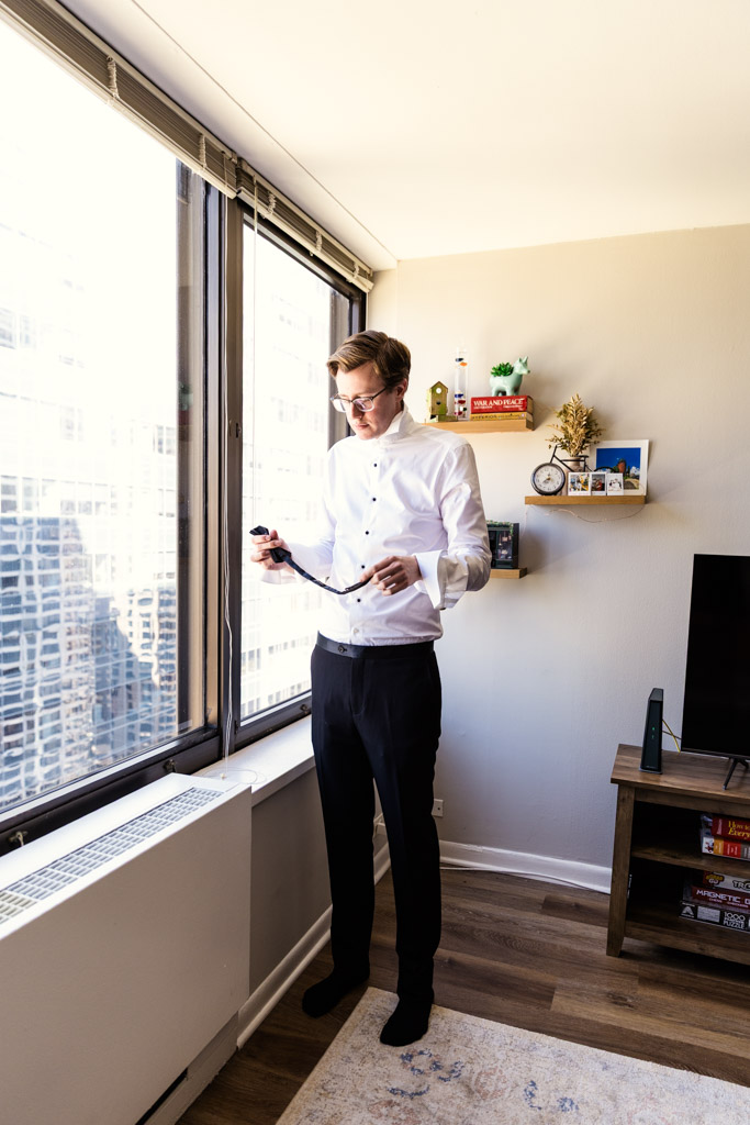 Groom in formal wear stands by a window in an apartment, preparing to put on a tie before his Salvatore's wedding celebration