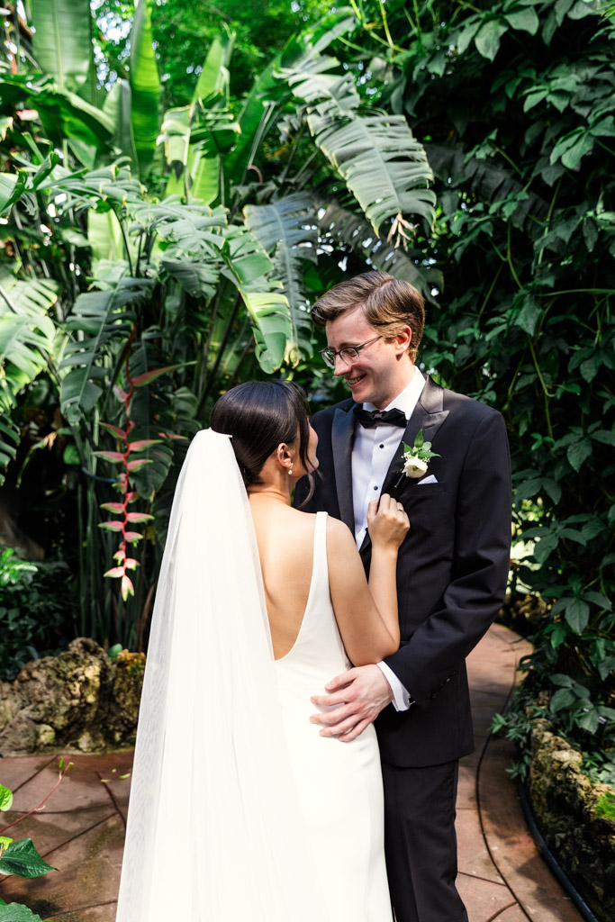 A bride and groom smile at each other while standing on a garden path surrounded by lush green plants inside Lincoln Park Conservatory