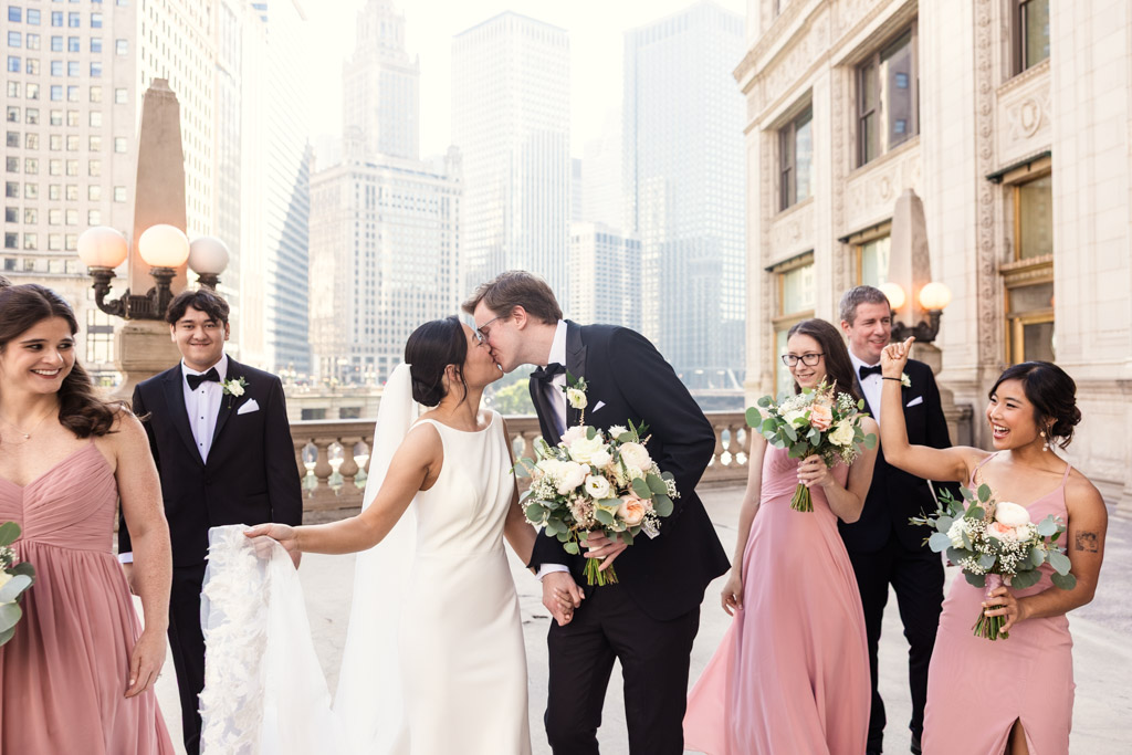 Bride and groom kiss surrounded by happy bridesmaids and groomsmen near the Wrigley Building in downtown Chicago