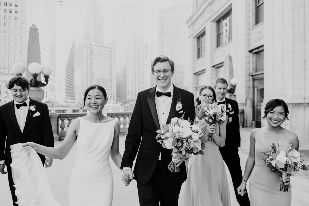 Black and white photo of happy bride and groom walking near the Wrigley Building in downtown Chicago with their wedding party in formal attire
